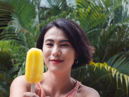 Woman eating ice cream. Happy beautiful Asian woman wearing casual pink tank top holding yellow ice cream on green tropical palm leaves background, outdoor. Smiling female enjoying ice lolly in summer.の写真素材