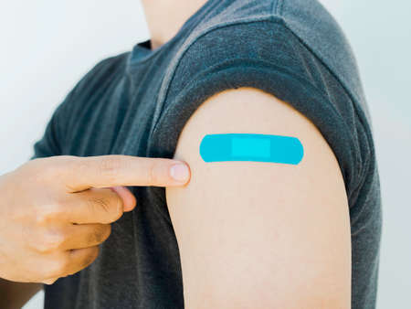 Vaccinations, bandage plaster on vaccinated people concept. Man hand pointing on blue adhesive bandage on own shoulder who wearing grey shirt after vaccination treatment on white background, close up.の写真素材