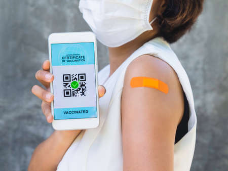 International certificate of vaccination, smart digital passport with QR code on smartphone screen showing by the vaccinated woman who wearing face mask and orange bandage plaster on his shoulder.の写真素材