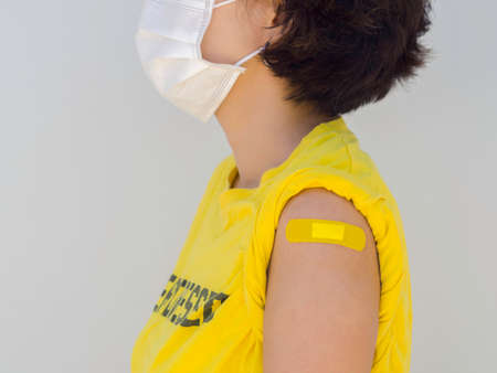 Vaccinations, bandage plaster on vaccinated women's arm concept. Yellow adhesive bandage on female shoulder who wearing yellow shirt, white face mask after vaccination treatment on white background.の写真素材
