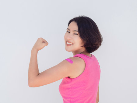 Vaccinations, bandage on vaccinated people's arm concept. Beautiful attractive Asian woman wearing pink tank top showing pink bandage and strong gesture with smile after vaccination treatment.の写真素材