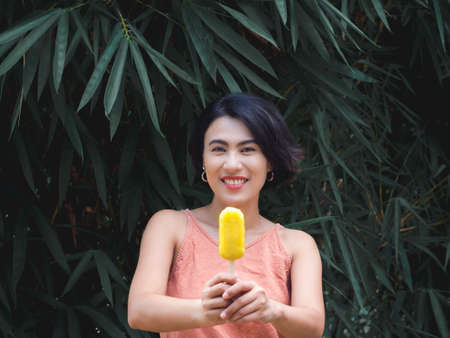 Woman eating ice pop. Happy beautiful Asian woman wearing casual pink tank top holding yellow ice pop on green tropical palm leaves background, outdoor. Smiling female enjoying ice lolly in summer.の写真素材