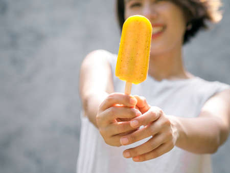 Close up yellow ice pop in beautiful happy Asian womanâ€™s hand wearing casual white sleeveless...