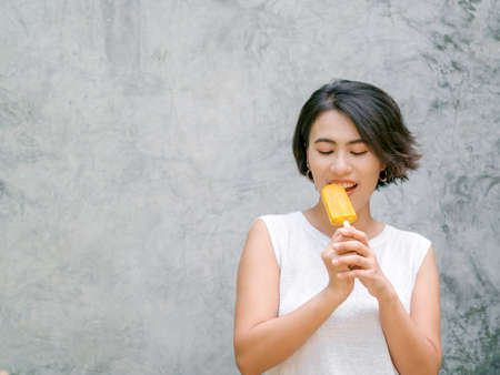 Woman eating ice pop . Happy beautiful Asian woman short hair wearing casual white sleeveless shirt holding yellow ice pop , outdoors with copy space. Smiling female enjoying ice lolly in summer.の写真素材
