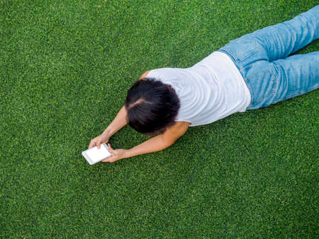 Beautiful happy Asian woman black short hair in white sleeveless shirt and blue jeans looking at smartphone with smiling while lying on green artificial grass, top view.の写真素材