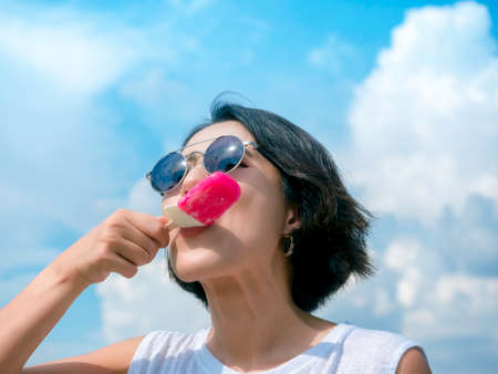 Happy beautiful Asian woman short hair wearing sunglasses and casual white sleeveless shirt holding pink ice cream on blue sky background in summer.の写真素材