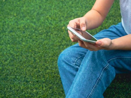 Close-up mobile phone in hands of woman wearing white sleeveless shirt and blue jeans sitting with relax on green artificial grass background with copy space. Hands holding a smartphone.の写真素材
