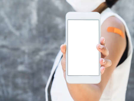 Mockup phone, white blank screen on smartphone holding and showing ing by the vaccinated woman who wearing white sleeveless blazer,  face mask and bandage plaster on his shoulder with copy space.の写真素材