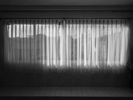 White transparent curtain at the window in the dark room. Large horizontal window with closed white fabric curtain.の写真素材