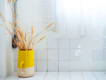 Yellow ceramic vase with dried rice plants on white clean grid tiles wall near the window with white transparent fabric curtain with copy space.の写真素材