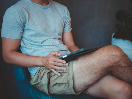 Hand man using digital tablet. A young person, man in casual grey shirt and shorts sitting and...