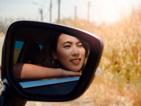 Happy Asian woman with short hair traveling by car. Attractive female travelers enjoy and smiling while looking at the view outside with sunshine, summertime, view from side view mirror.の写真素材