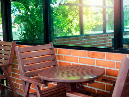 Empty red cozy wooden chairs and round wood coffee table near the glass window and brick wall with green garden in the cafe, summertime.の写真素材