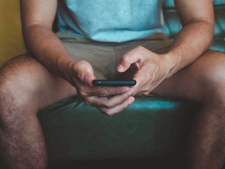 Hand man holding smartphone. A young person, man in casual grey shirt and shorts sitting and...