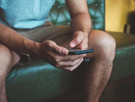 Hand man using smartphone. A young person, man in casual grey shirt and shorts sitting and interacting with technology at home. Relaxation businessman working online at anywhere concept.の写真素材