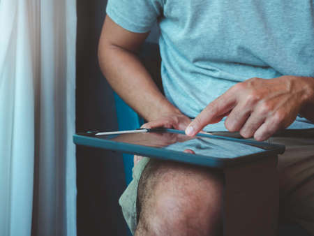 Hand man using digital tablet. A young person, man in casual grey shirt and shorts sitting and interacting with technology at home. Relaxation businessman working online at anywhere concept.の写真素材