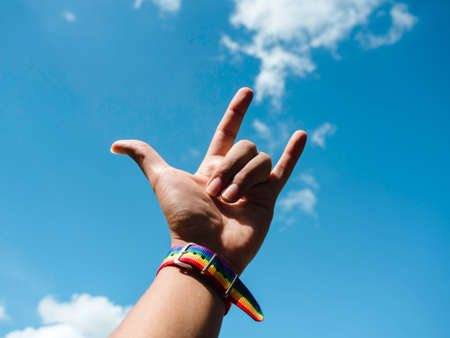 Small rainbow flag belt on gay people wrist with I LOVE YOU finger sign gesturing up into the blue sky background. LGBT concept with pride colors and rainbow flag strip.の写真素材