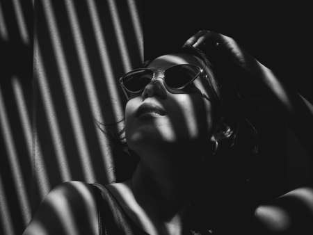 Black and white style female portrait. Beauty Asian woman short hair wearing sunglasses and a grey tank top with strips of light and shadows on her face, shadows from the slatted roof.の写真素材