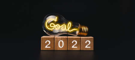 2022 Happy New year banner with goal target and successful concept. The 2022 numbers on wooden cube blocks and a light bulb with shining neon light words on dark background, modern and minimal style.の写真素材