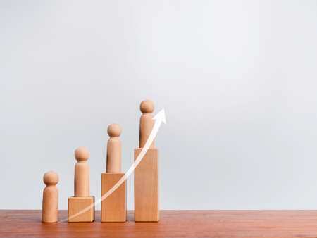 Population growth concept. Rising up arrow and wooden figures standing on a growing graph chart steps arranged by wood cube blocks on wood table and white background with copy space.の写真素材