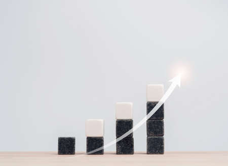 Growth and success business chart built with black and white blocks as steps with a rising arrow on wood table, white background. Increase revenue, benefit, profit, and economic improvement concept.の写真素材