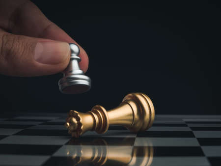 Close-up little silver pawn chess piece wins golden queen and falls on chessboard, dark background....
