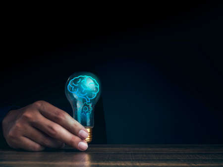 Creative, idea, innovation, knowledge, and inspiration concept. The human brain icon is glowing inside the digital light bulb in man's hand on the wooden table and dark background with copy space.の写真素材