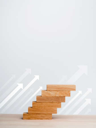 White rise up arrows with wooden blocks arranged as a steps chart on a wooden desk and clean white background with copy space, vertical style. Business growth process and economic improvement concept.の写真素材