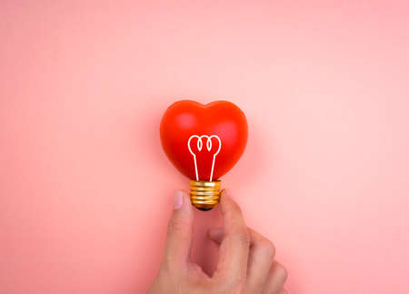 Hand holding light bulb icon symbol on red heart ball on pink pastel background, minimal style. Love, care, sharing, giving, wellbeing, inspiration, and idea concepts.の写真素材