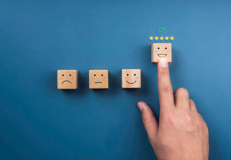 Customer service evaluation, ratings, feedback, client experience, and satisfaction survey concepts. Checkmark at five stars and hand pointing at happy face on wooden cube block on blue background.の写真素材