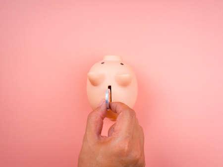 Pink piggy bank on pink pastel background while hand putting a silver coin, top view, minimal style....