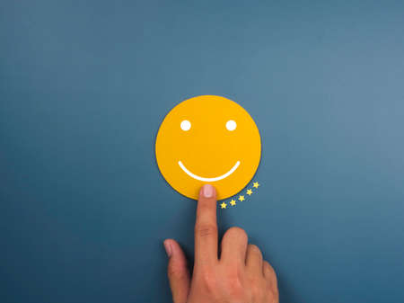 Customer service evaluation, feedback, client experience, satisfaction survey concepts. Happy smile on yellow face chosen by customer's hand with five stars ratings on blue background, minimal style.の写真素材
