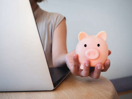 Money box on hand. Hand holding the pink piggy bank while using laptop computer on desk. Saving money wealth and financial concept, Business, online banking, finance, investment, financial planning.の写真素材