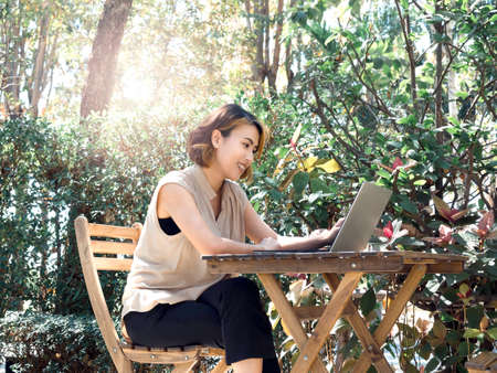 Happy smile Asian woman short hair in beige sleeveless shirt sit and working with laptop computer on wood table, relaxing at the outdoor, surrounded with green plants in the garden. Work everywhere.の写真素材