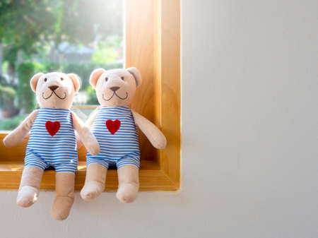 Two happy smiling brown bears, dolls in blue and white striped shirts with red hearts, sitting on the edge of wooden glass window on white wall background with copy space. Love, lover, couple concept.の写真素材
