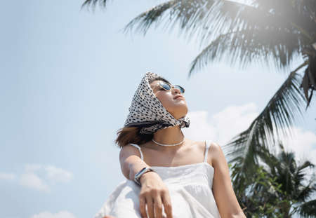 Beautiful happy Asian woman portrait in casual white shirt wearing sunglasses and hair scarf, looking up to the sunshine at the beach under the coconut palm trees and blue sky background in summer.の写真素材