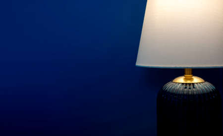 Close-up luxury table lamp with white fabric, blue glass, and gold color materials shining on dark blue wall background with copy space. Lighting lamp in the dark.の写真素材
