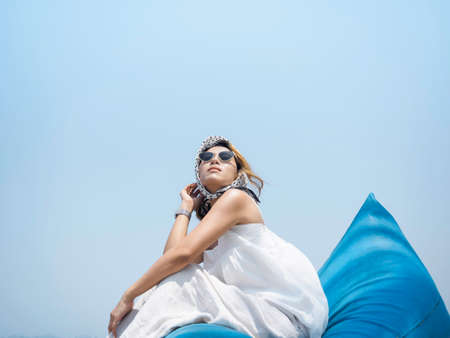 Attractive Asian woman in casual white shirt and trousers wearing sunglasses and hair scarf look up the sky while sitting on blue bean bag seat on blue sky background on sunny day in summer.の写真素材