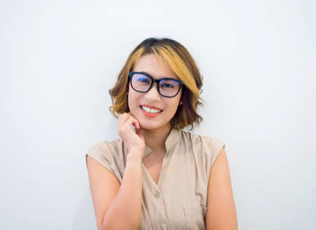 Happy smiling pretty Asian woman short hair in black eyeglasses and beige sleeveless shirt posing with a hand on chin, looking at camera on white background. Working female portrait with glasses.の写真素材