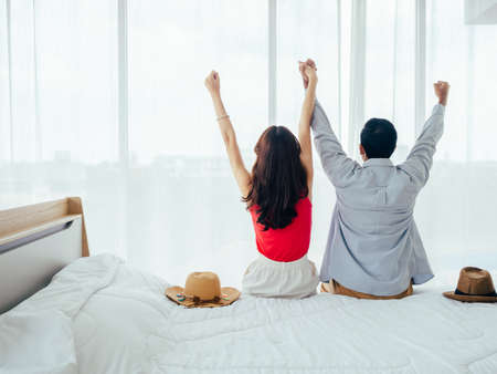 Couple of tourists, happy holiday. Summer vacation. Portrait of back view of young Asian man and woman raising hands with happy and joyful on white bed near curtain at the window in hotel room.の写真素材