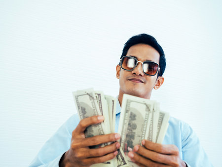 Portrait of happy, rich Asian young businessman in casual blue shirt and wearing sunglasses enjoying while holding and counting dollars money banknotes on white wall background.の写真素材