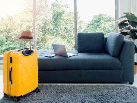 Empty dark sofa seat with passport, laptop computer, and beach hat on yellow travel suitcase on the carpet near the huge glass window at home. Ready to travel, happy holiday, summer vacation concept.の写真素材
