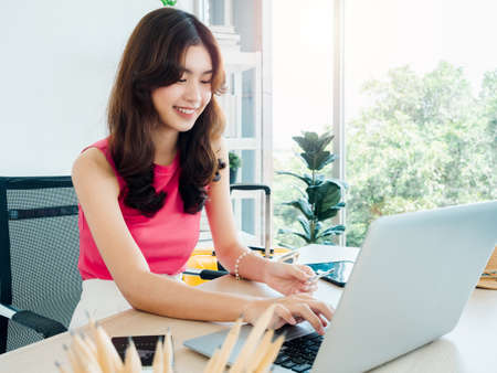 Happy young pretty Asian woman holding credit card while using laptop computer for flight booking, hotel room for summer trip at home, ready to travel, happy holiday, summer vacation concept.の写真素材
