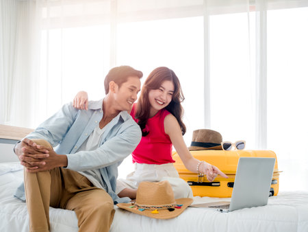 Preparing trip and vacation plan concepts. Young pretty Asian woman and man in denim shirt looking at the laptop and map on the bed with suitcase in bedroom. Travel planning on summer holiday.の写真素材
