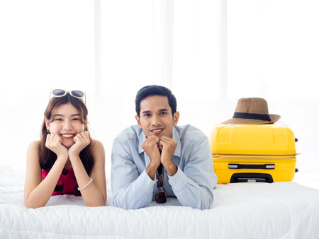 Happy couple travelers on honeymoon trip. Asian man in denim shirt and young beautiful woman smile and lying together with yellow suitcases on bed in bedroom. Happy holiday. Colorful summer vacation.の写真素材