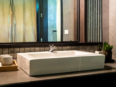 Wet stainless steel faucet on white ceramic sink basin in clean white bathroom interior with ceramic cups, liquid soap pump bottle near the mirror. Modern contemporary style design of bathroom.の写真素材