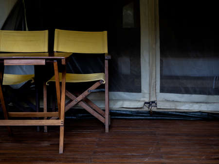 Two empty camping wooden chairs with yellow fabric seats and backrest and wood table on the terrace in front of the room of large tent. Folding outdoor chairs on white glamping tent.の写真素材