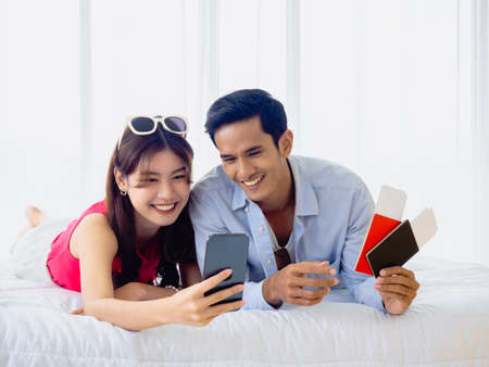 Happy Asian couple with flight booking for summer trip, travel abroad. Young man and woman in casual holding and looking at smartphone screen and holding passport and ticket while lying on bed.の写真素材