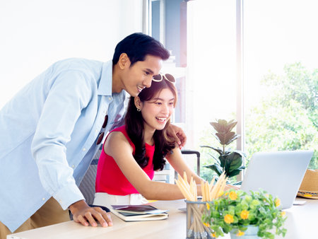 Happy Asian couple, young woman and man using laptop computer on desk together for flight booking, hotel room and trip information in office, ready to travel, happy holiday, summer vacation concept.の写真素材