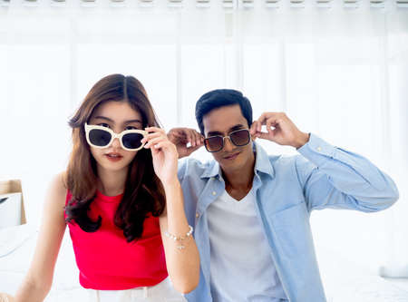Happy Asian couple in summer, young man and woman wearing sunglasses and touching it and looking at camera on bed in the bright bedroom near the glass window, holiday travel, summer vacation concept.の写真素材